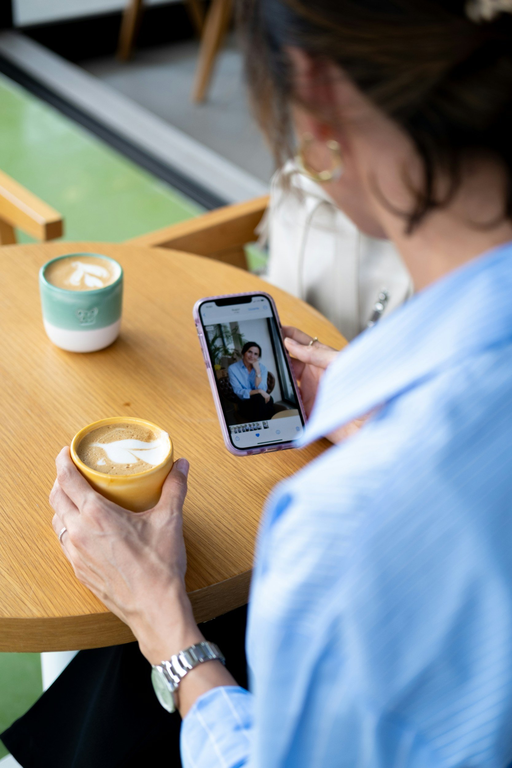 A woman sitting at a wooden table takes a photo of herself with a smartphone while holding a cup of coffee. There is another cup of coffee on the table.