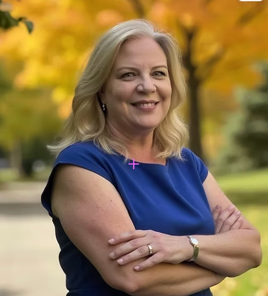 A woman with blonde hair wearing a blue dress, smiling with arms crossed, outdoors in a park with autumn-colored trees.