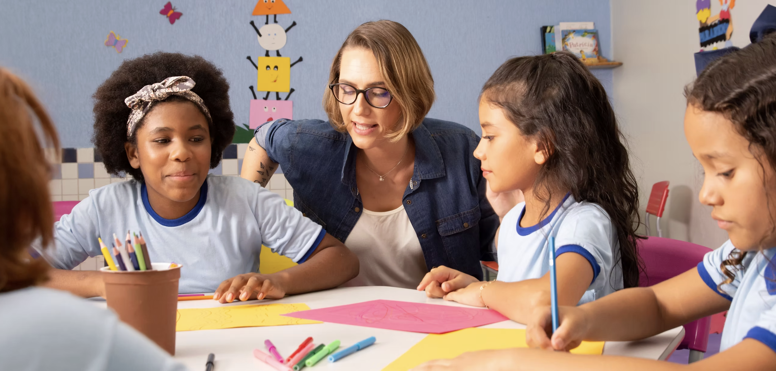 A teacher in a denim shirt teaches a group of young girls in school uniforms at a table, surrounded by art supplies and colorful wall decorations.