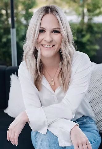A woman with shoulder-length blonde hair sitting outdoors, smiling, wearing a white blouse and a necklace.