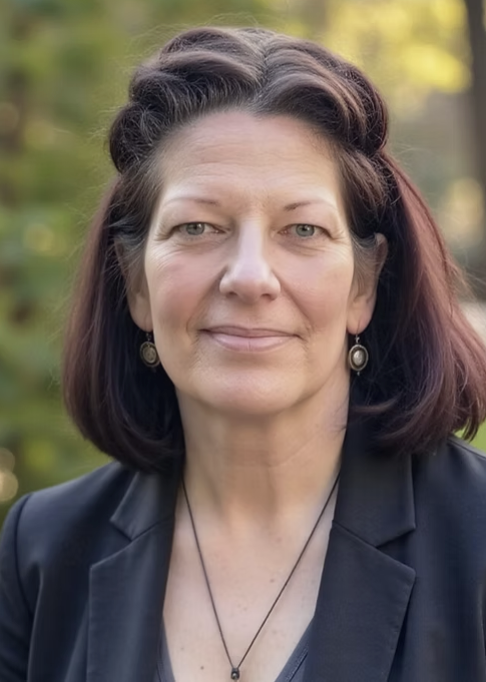 Portrait of a woman with shoulder-length dark hair, wearing a black blazer and earrings, outdoors with blurred green foliage in the background.