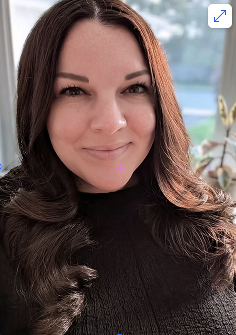 A woman with long, dark brown hair smiling indoors near a window with plants and a butterfly decoration in the background.