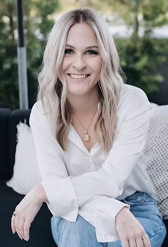 A smiling woman with blonde hair wearing a white blouse and blue jeans, sitting outdoors with trees in the background.