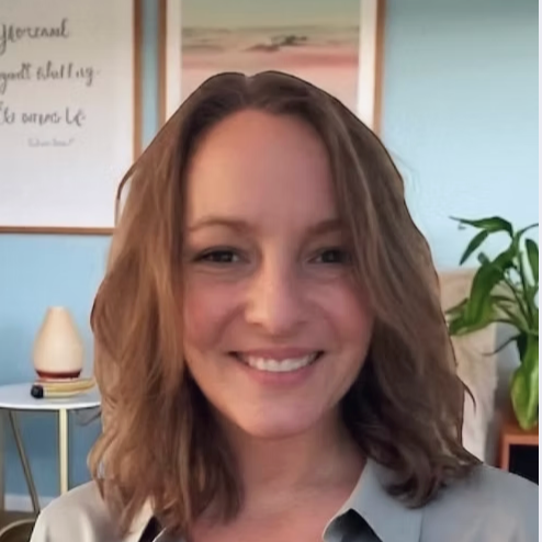 A woman with shoulder-length wavy hair smiling in a room with blue walls, a framed picture, a white lamp, and a potted plant in the background.