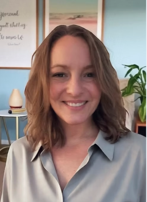 A young woman with shoulder-length wavy brown hair smiling indoors with a light blue wall, a framed picture of a beach, a white lamp, and a green plant in the background.