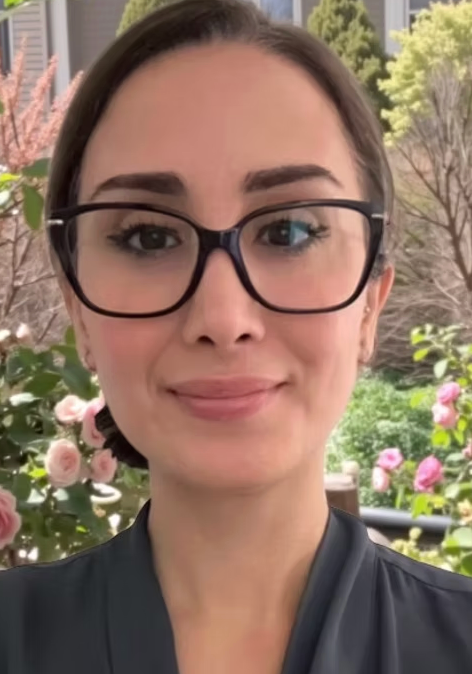 Portrait of a woman with glasses and dark hair, standing outdoors with flowering bushes and trees in the background.