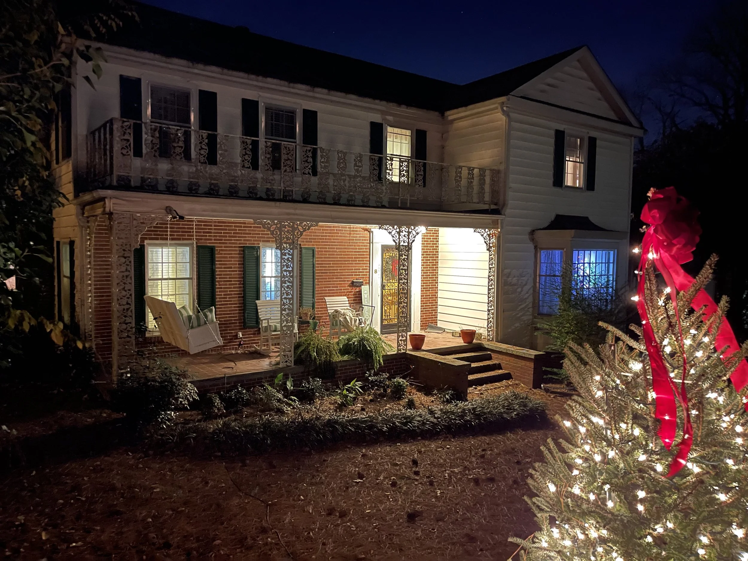 A two-story brick and white clapboard house with ornate ironwork porches, lit warmly at night, with a Christmas tree wrapped in lights and a red bow in the foreground.