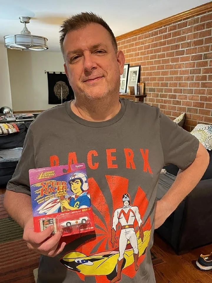 A man in a Racer X t-shirt smiling and holding a toy Mach 5 car from Speed Racer, standing in a living room.
