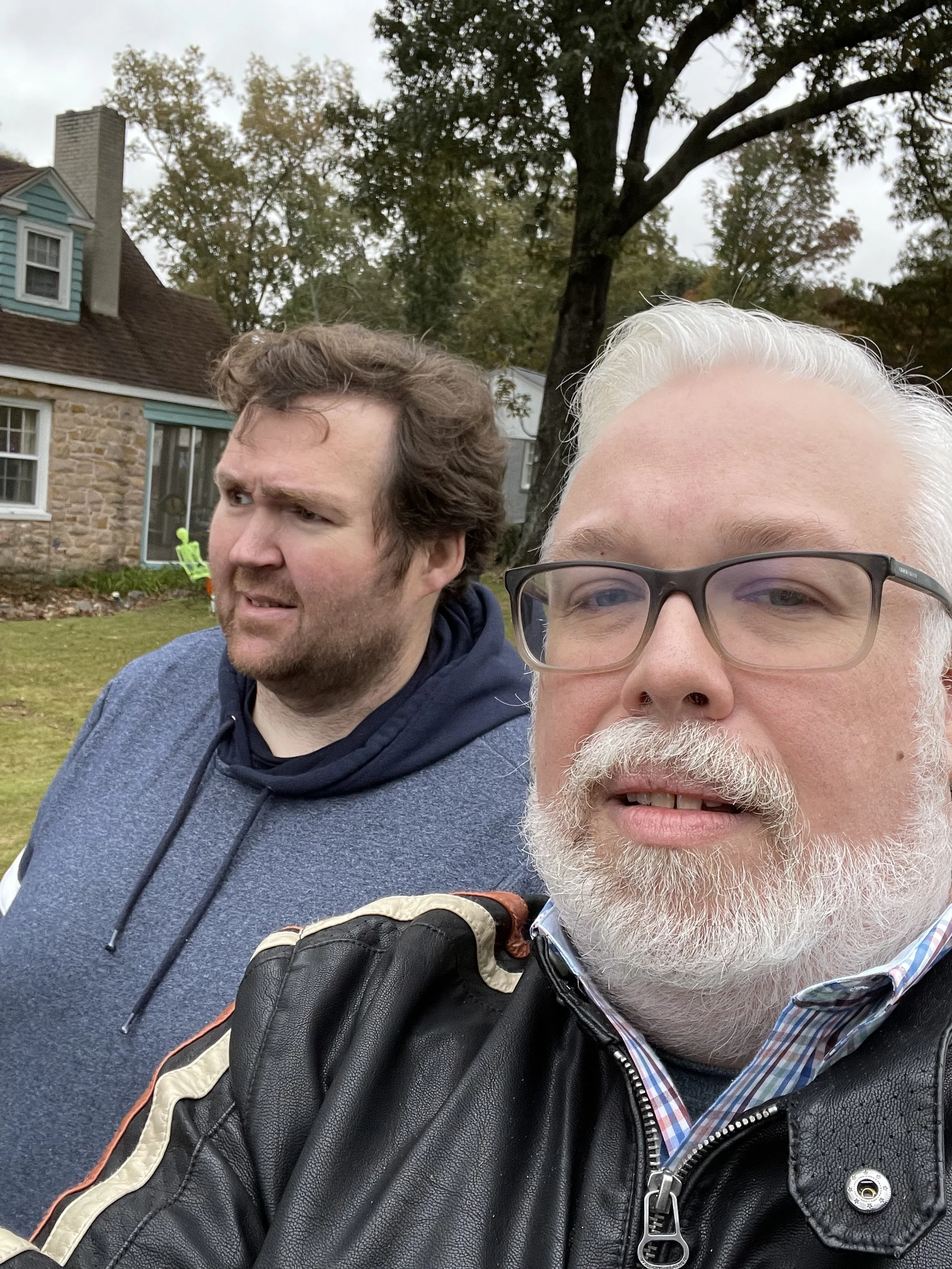 Two men taking a selfie on an overcast autumn day, walking through a residential neighborhood with a stone house and large trees behind them.