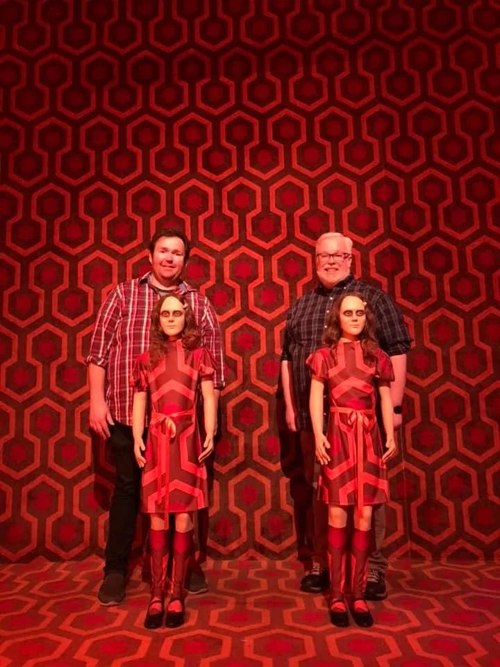 Two men standing with life-size figures of the Grady twins from The Shining, against a room covered in the iconic red hexagonal carpet pattern.