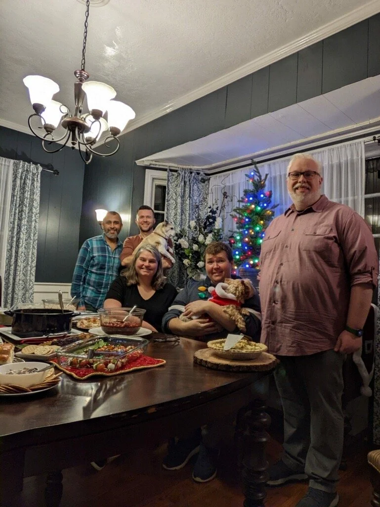 A group of five friends gathered around a dining table covered with food, a Christmas tree glowing in the background, one person holding a small dog in a holiday outfit.