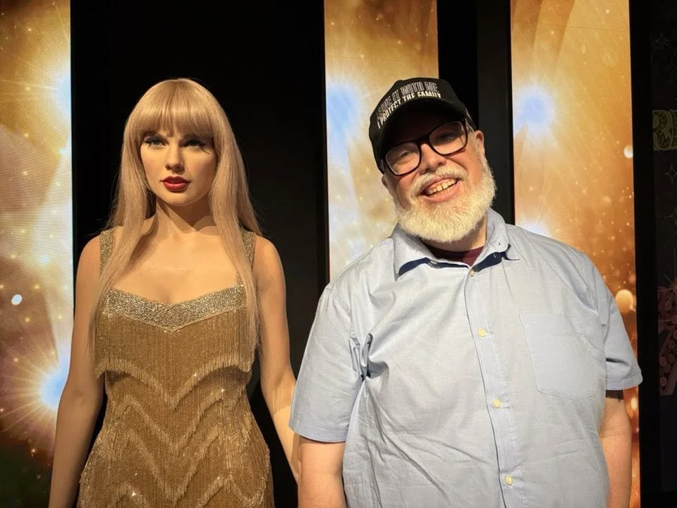 A bearded man in a blue shirt and black cap smiling next to a wax figure in a gold sequined dress at Madame Tussauds museum.