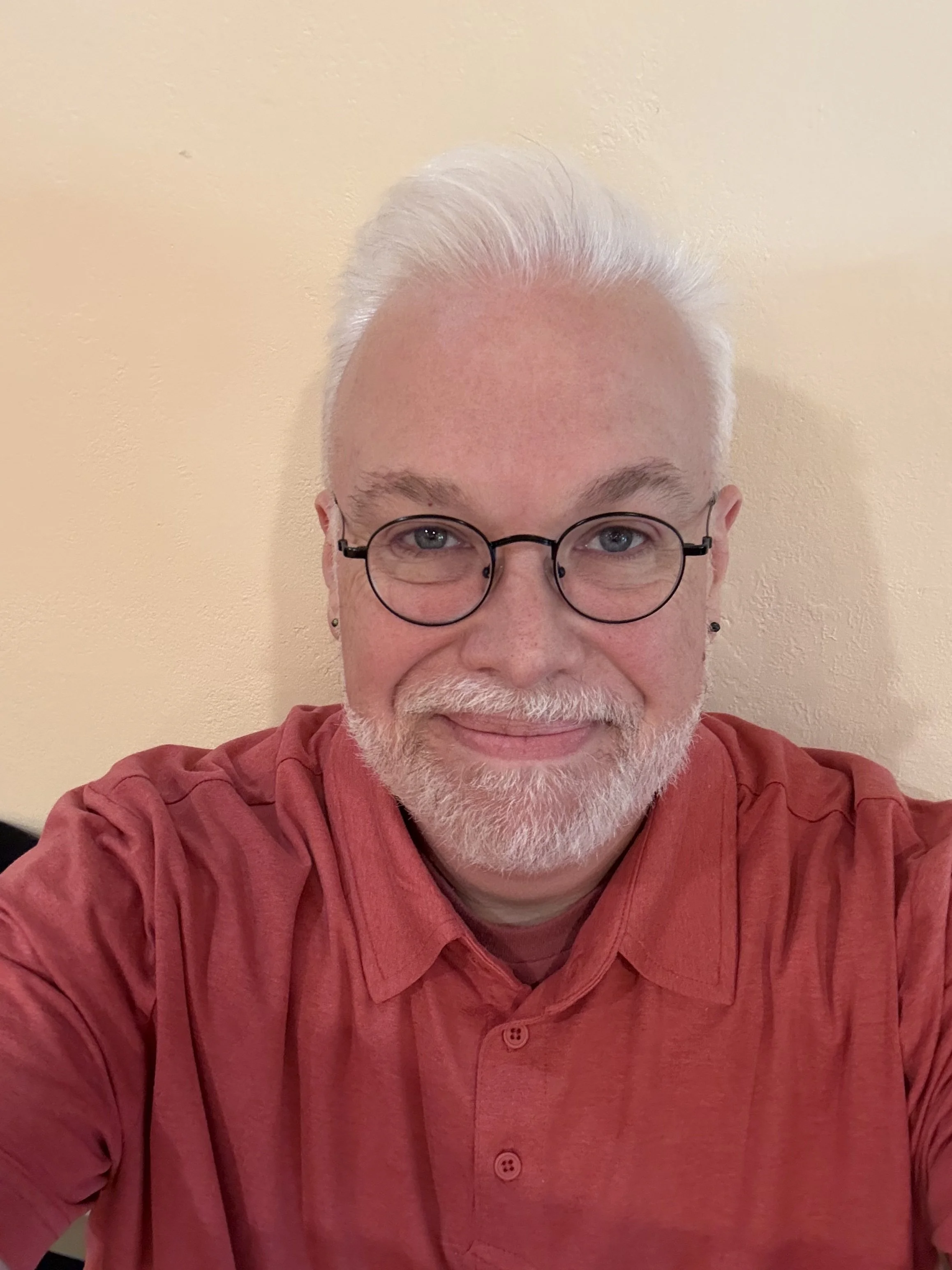 Jamie McDaniel smiling in a coral button-up shirt, with white hair swept back, a short white beard, round black-framed glasses, and small stud black opal earrings.