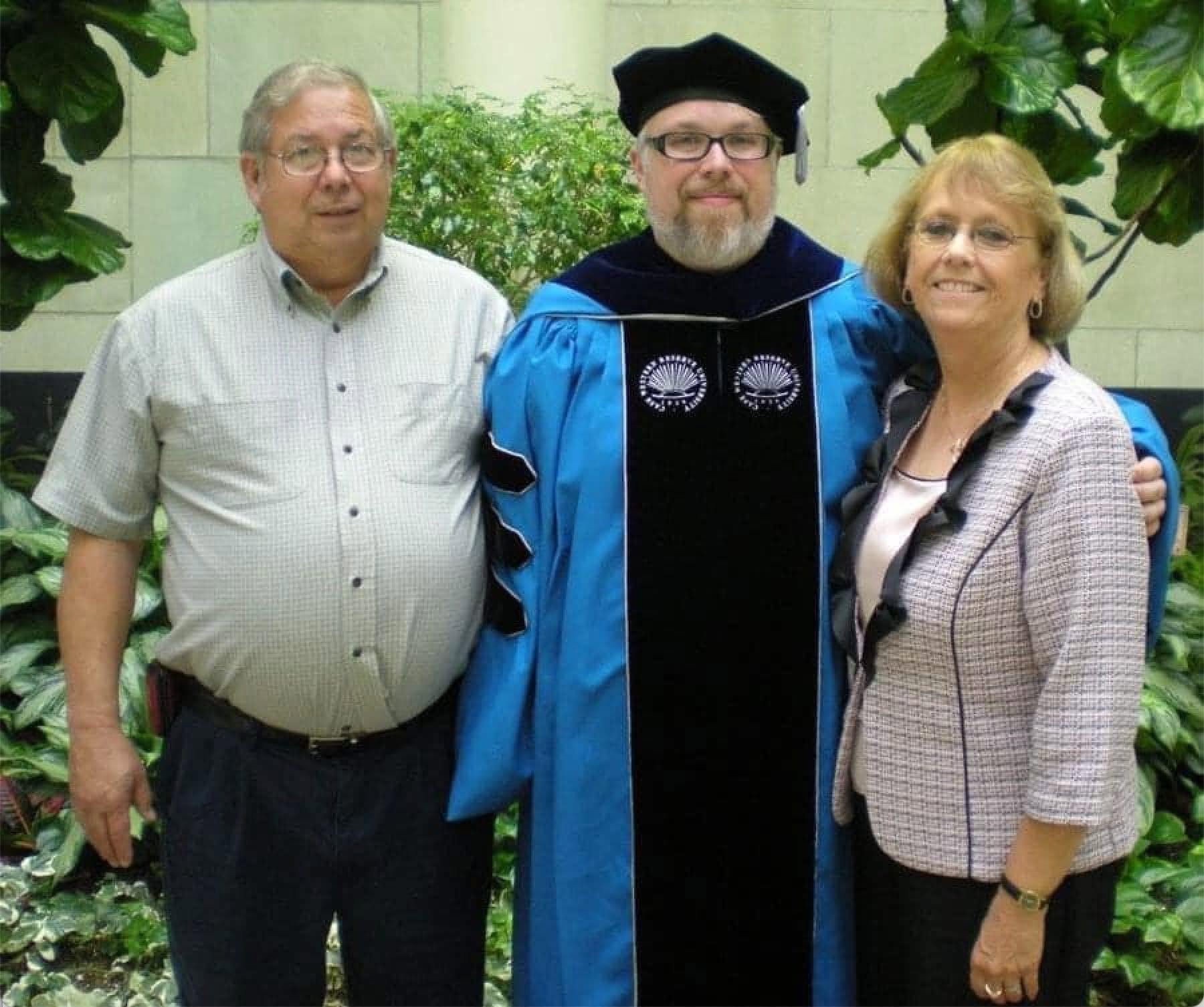 A man in blue doctoral regalia and cap standing between his parents outdoors at a university graduation ceremony.