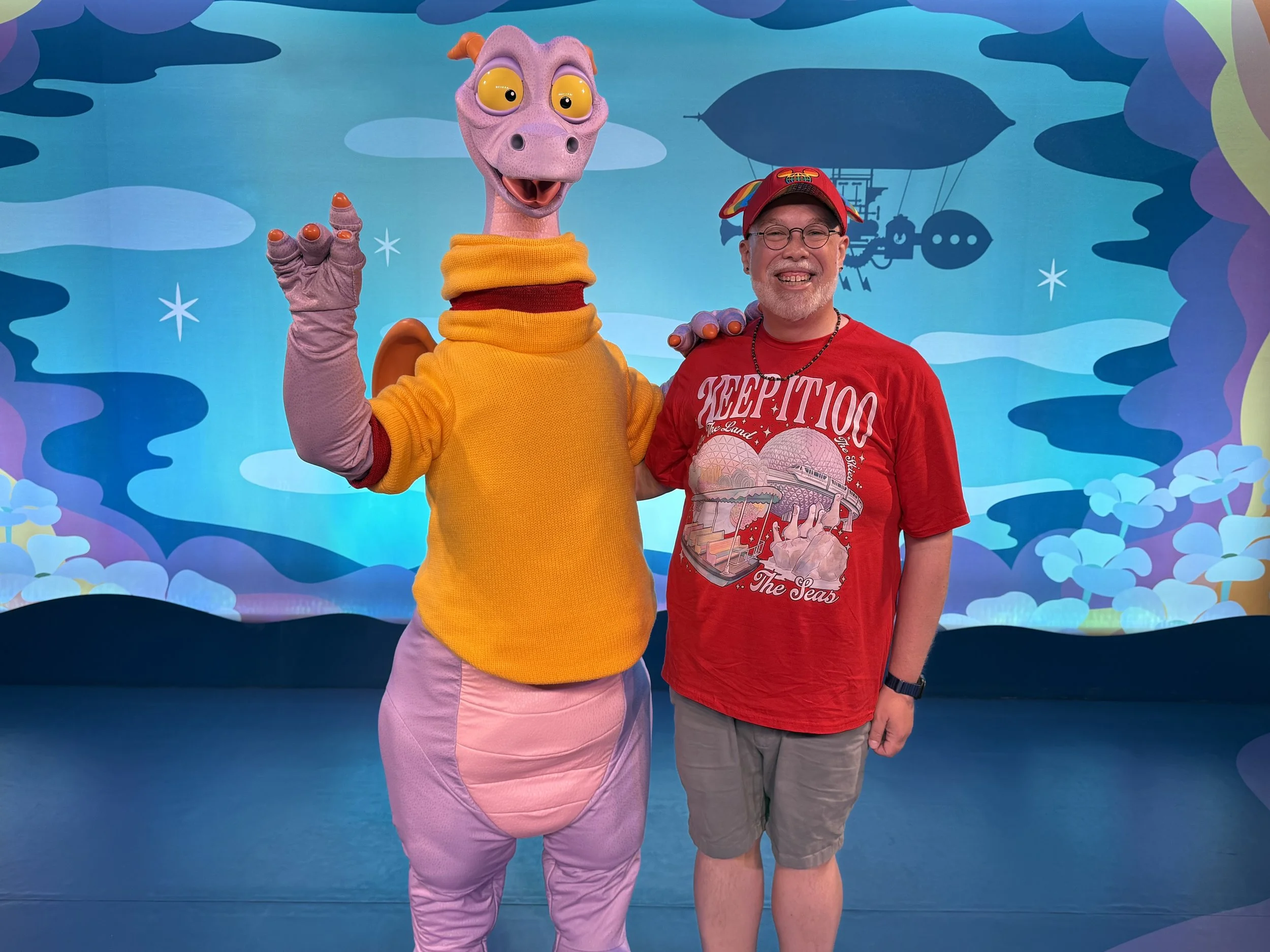 Jamie McDaniel smiling next to the character Figment at EPCOT, wearing a red Keep It 100 EPCOT attractions t-shirt against a colorful blue and purple sky backdrop.