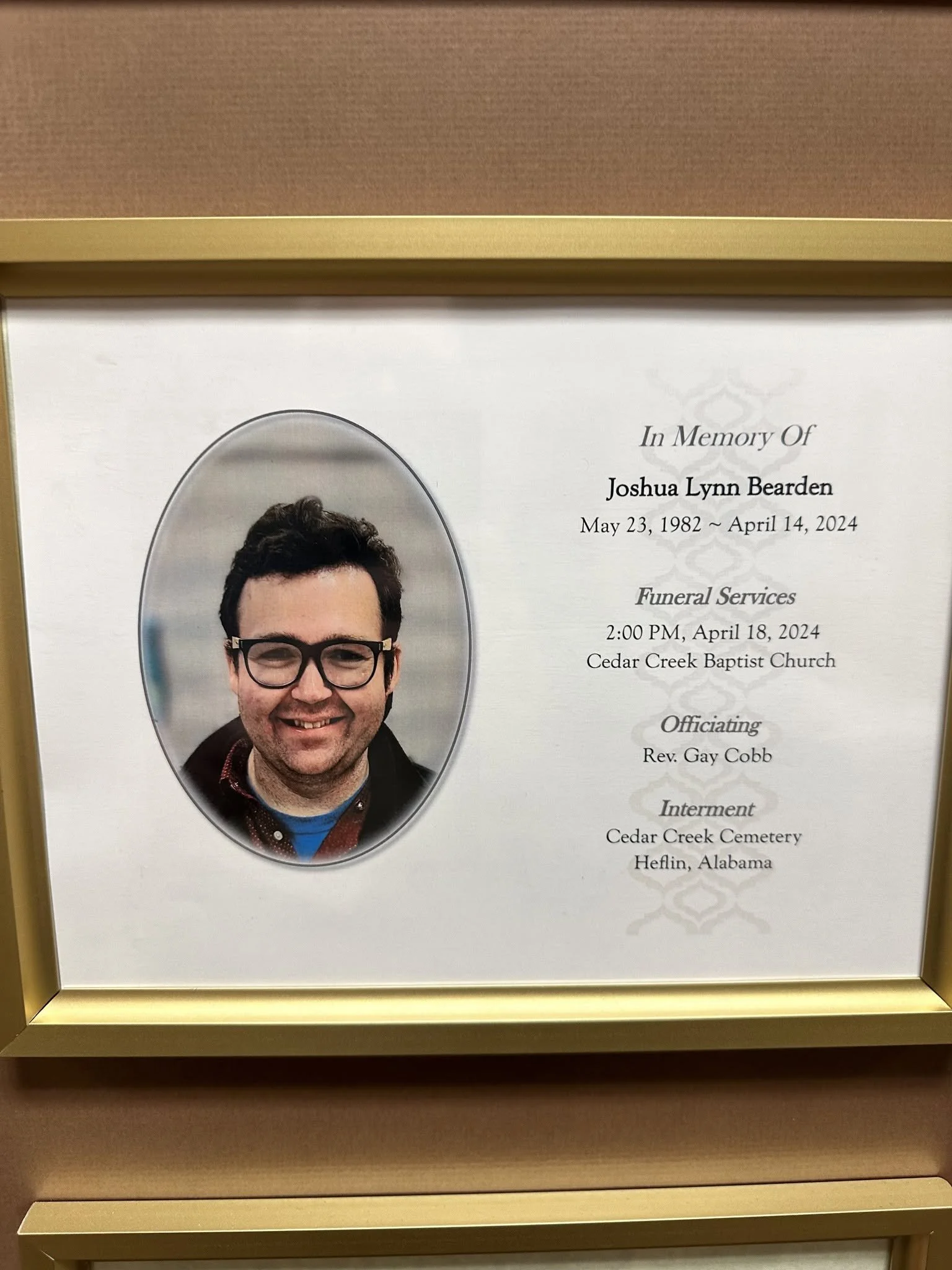 A framed funeral program with a portrait photograph of a smiling young man in glasses, reading In Memory Of Joshua Lynn Bearden.