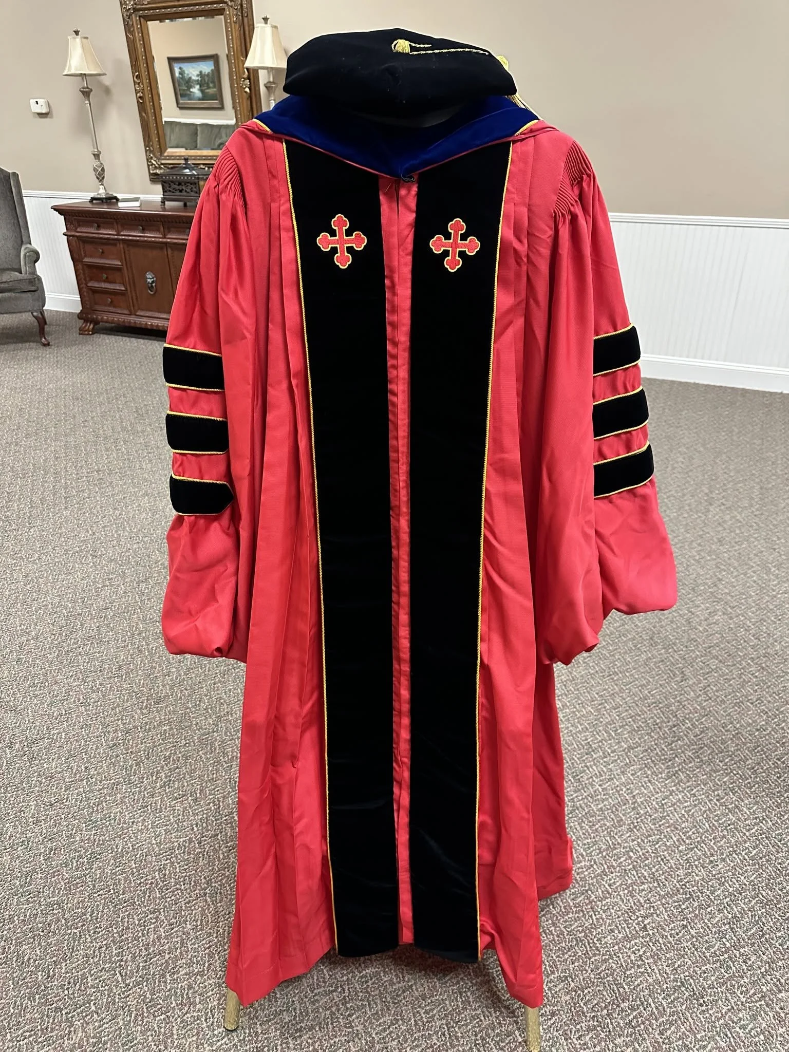 A red and black doctoral graduation robe with gold trim, cross emblems, and a blue-hooded cap displayed on a stand in a living room.
