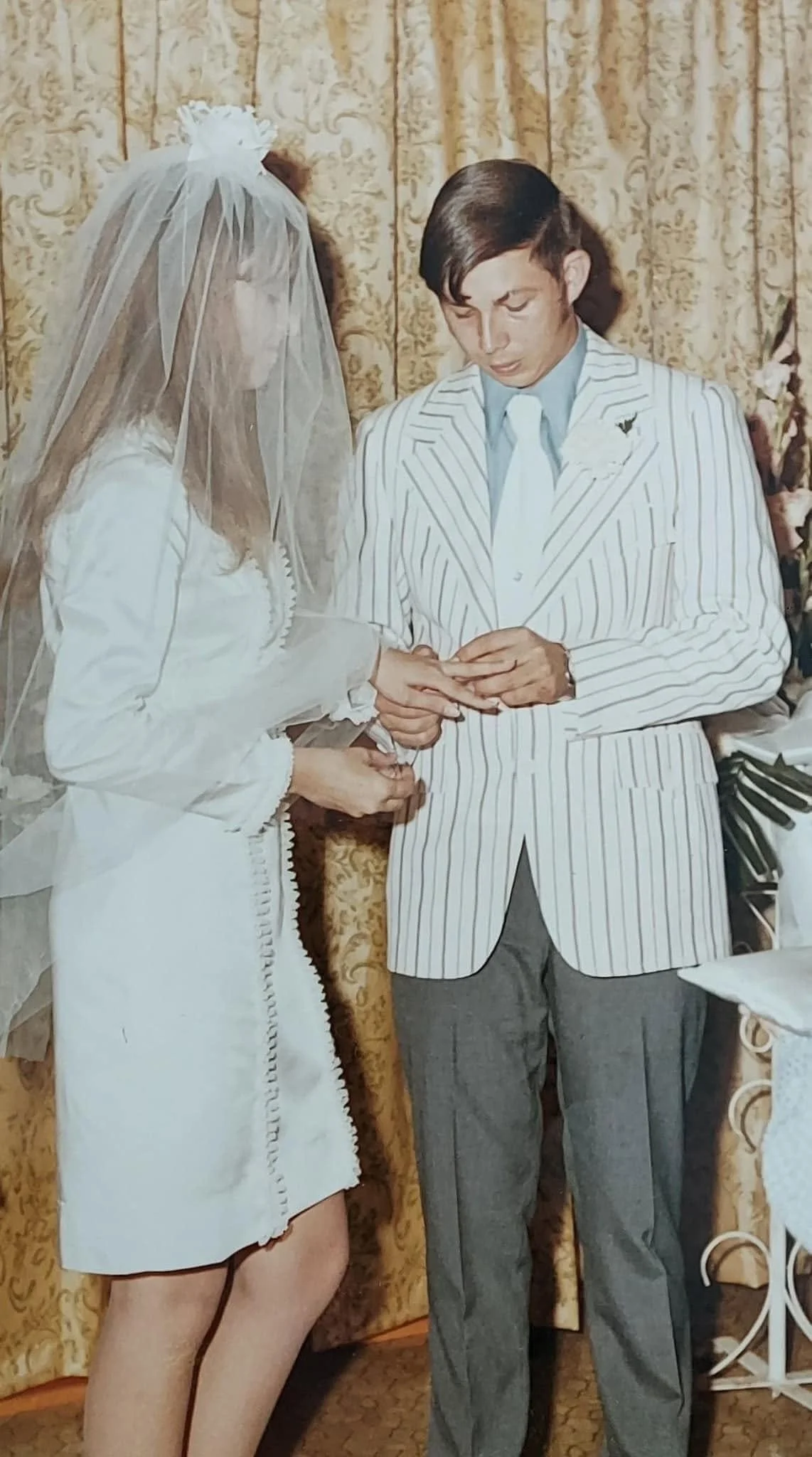A vintage color photograph of a young couple exchanging rings at their wedding ceremony, the bride in a short white dress and veil, the groom in a striped blazer.