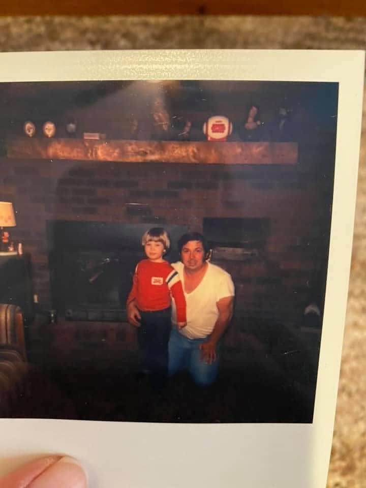 A Polaroid photograph of a young child in a red shirt standing with his father in front of a brick fireplace.