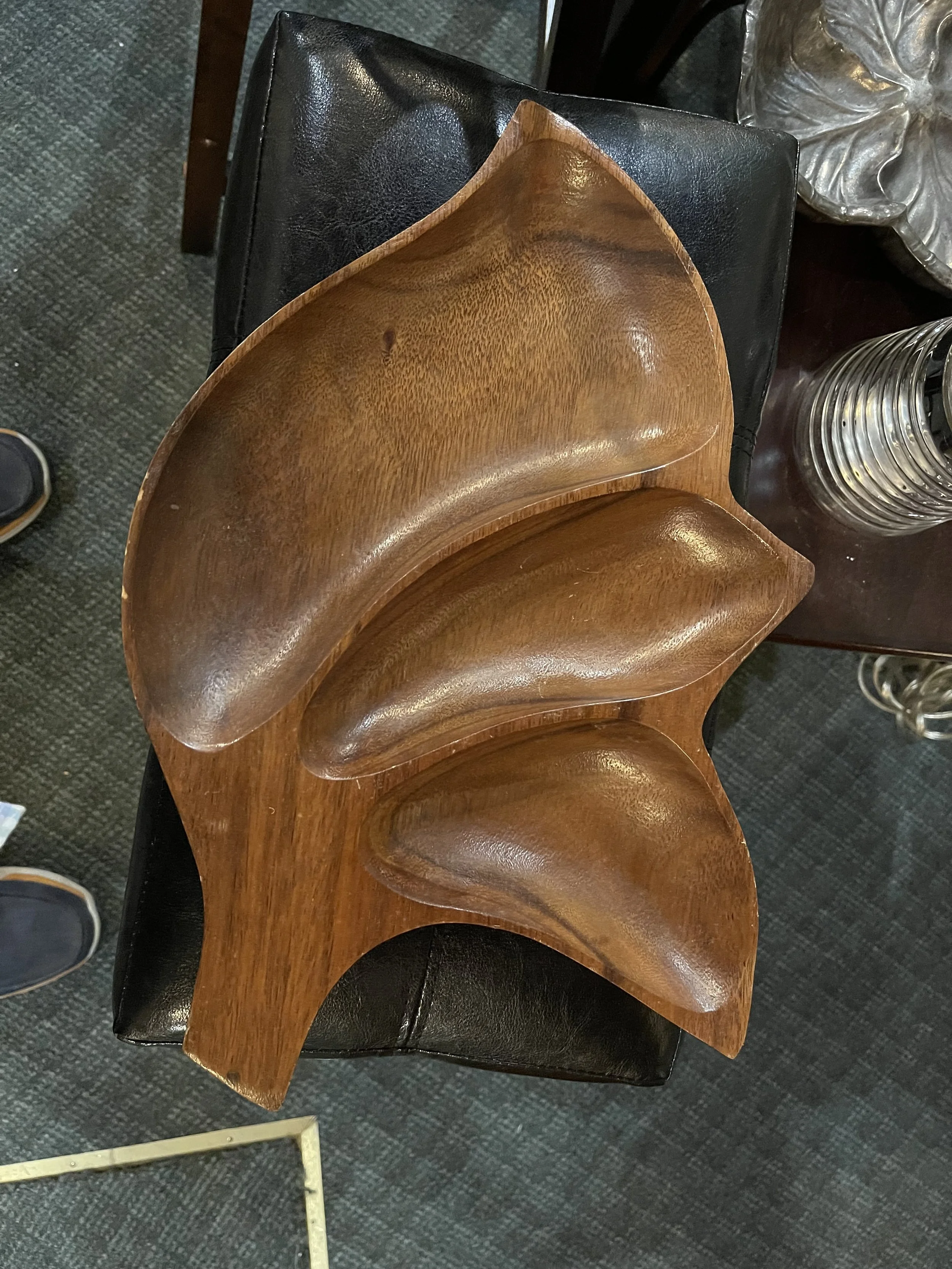 A carved wooden serving dish with organic, leaf-like curves in warm brown wood, resting on a black leather chair.