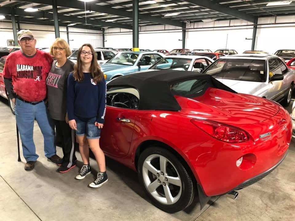 A man in an Alabama Crimson Tide shirt, a woman, and a teenage girl standing next to a red Pontiac Solstice convertible in a large car warehouse.