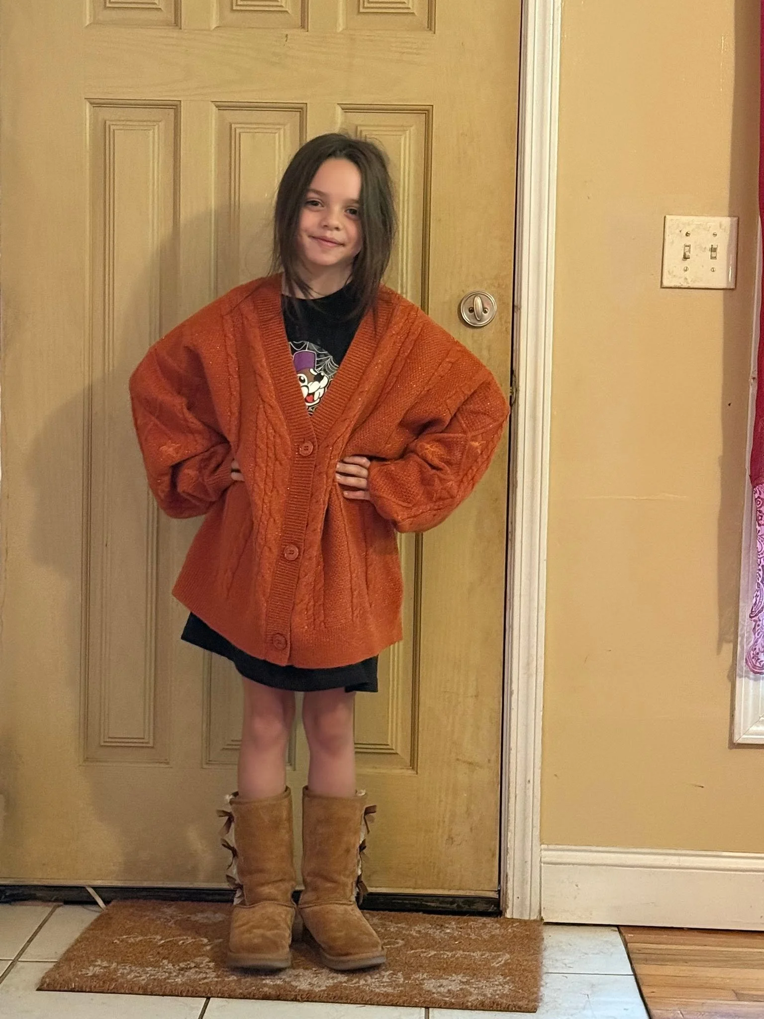 A young girl posing confidently in an oversized orange cable-knit cardigan, black dress, and brown boots, standing by a front door.