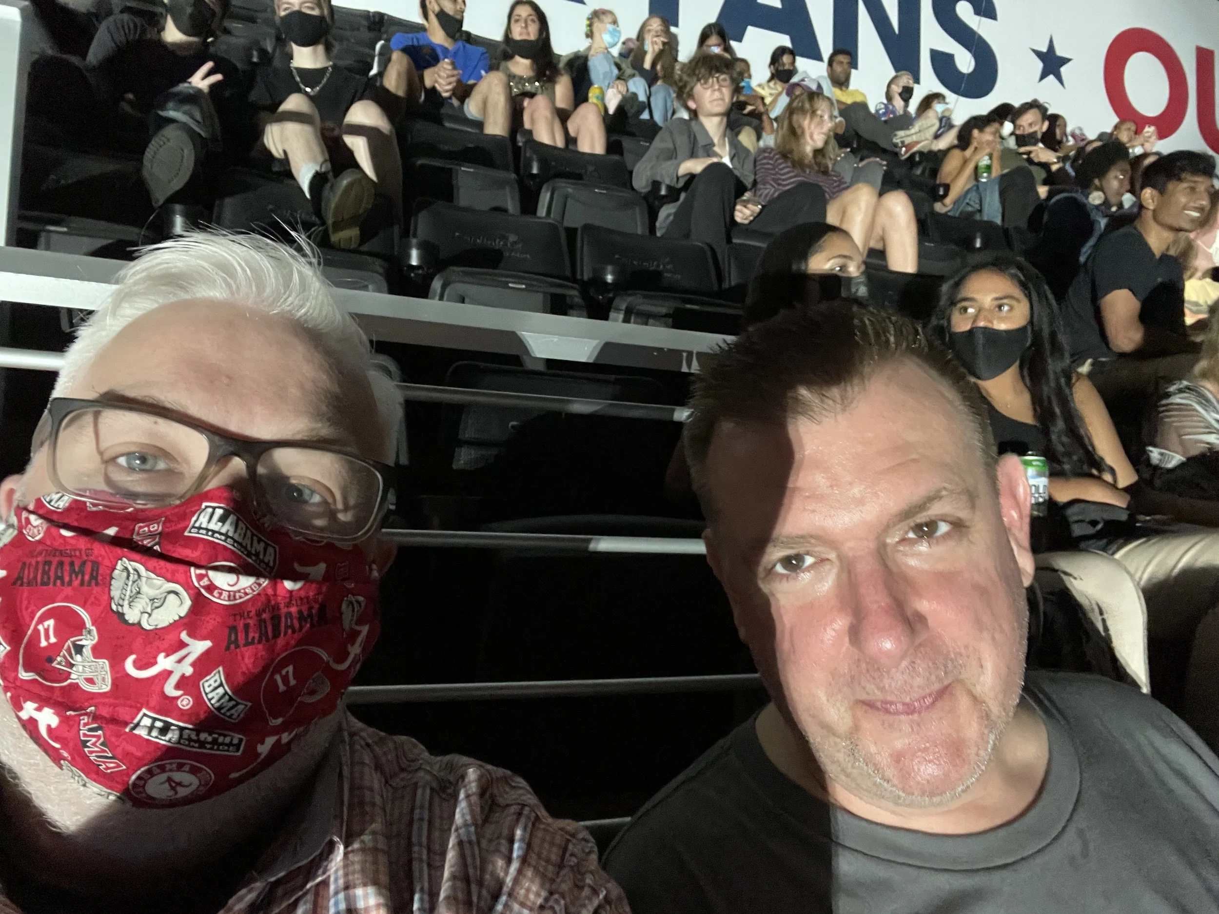Two men taking a selfie at a concert venue, one wearing an Alabama Crimson Tide face mask and plaid shirt, the other in a dark t-shirt, with arena seating and other concertgoers behind them.