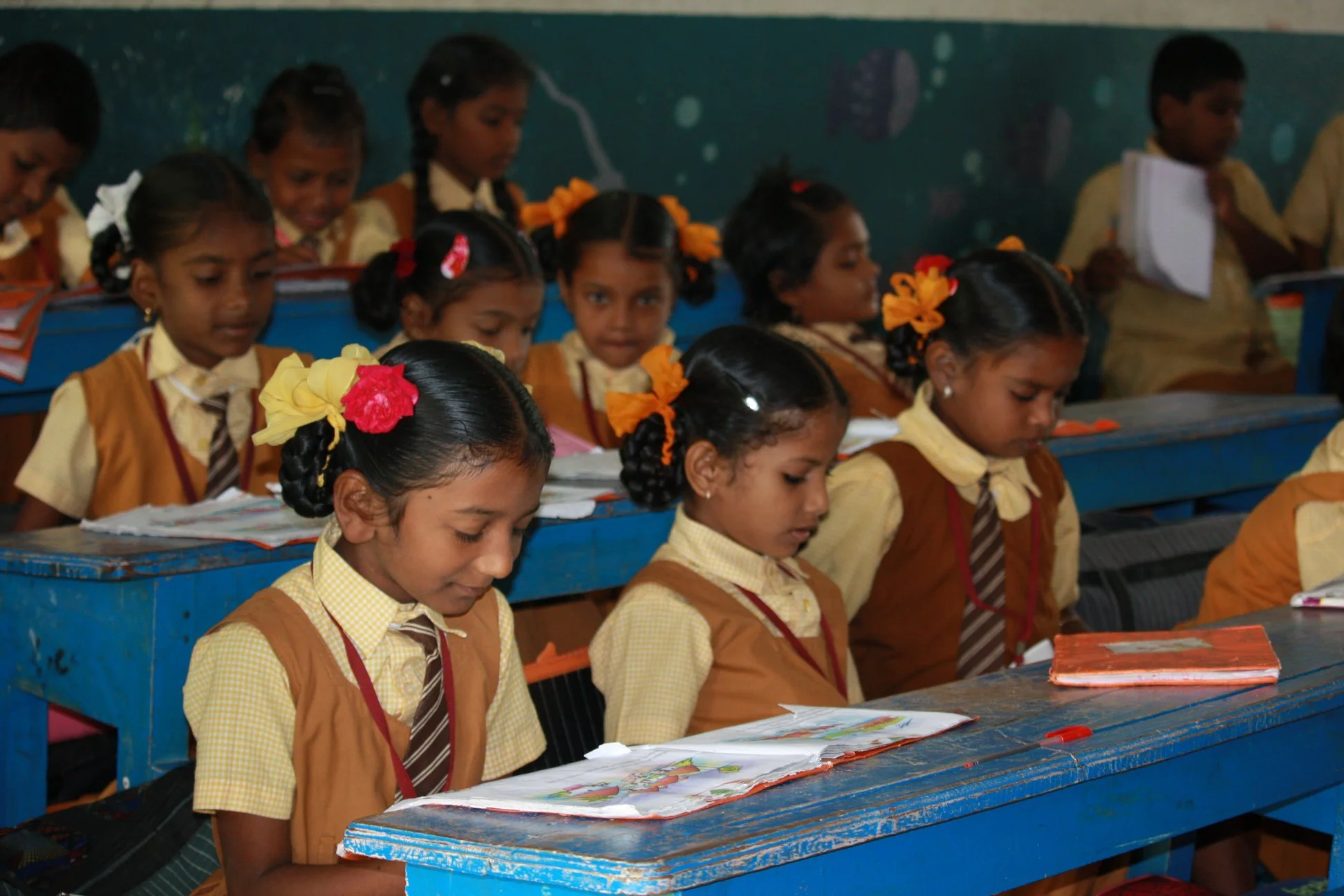 School classroom with young students in tan uniforms, sitting at blue desks, reading books.