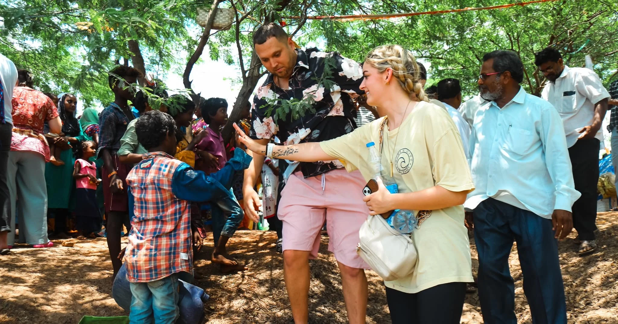 A group of people, including two women and several children, gathered outdoors under a tree. The women are engaging with the children, one with a high-five.