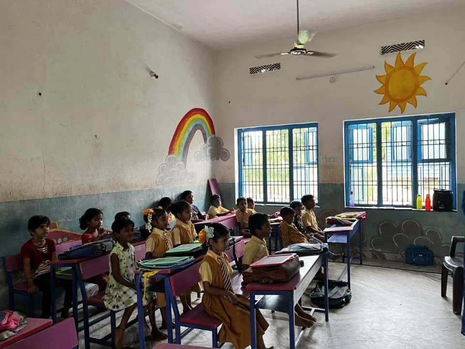 Classroom with students sitting at desks, windows, and colorful wall decorations including a rainbow, sun, and clouds.