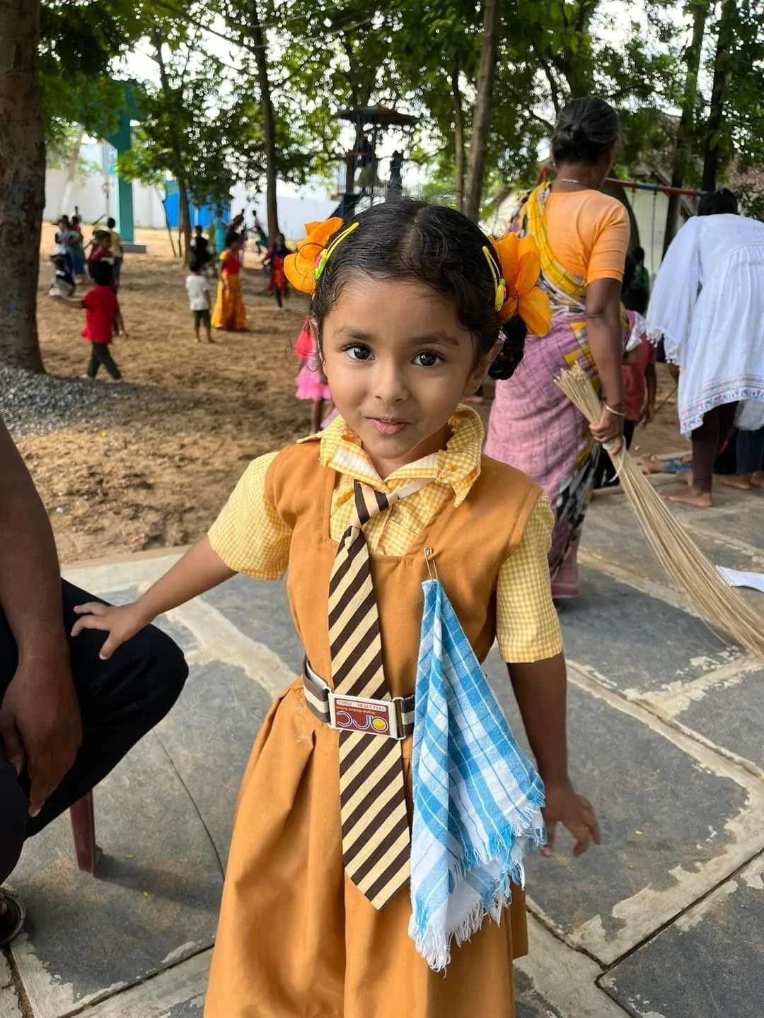 A young girl in a yellow school uniform with a checkered shirt, striped tie, and blue scarf, standing outdoors with other children and adults in the background, under trees.