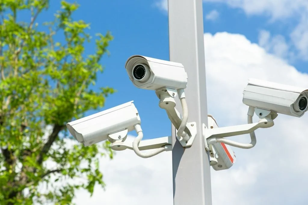Multiple security cameras mounted on a pole against a blue sky with clouds and a green tree in the background.