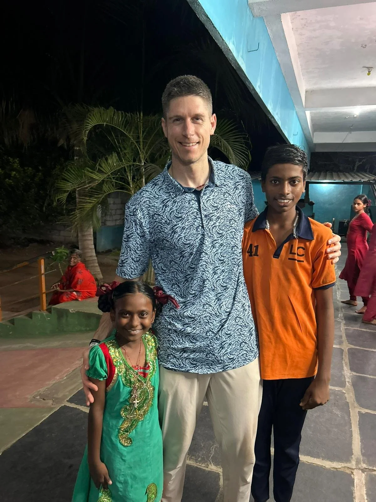 A man with two children, a girl and a boy, standing together outdoors at night, smiling at the camera. The girl is wearing a green dress with gold embroidery, and the boy is wearing an orange shirt with navy blue accents. In the background, there are other people and trees.