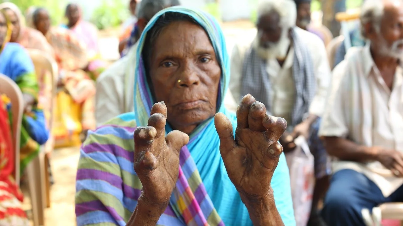 An elderly woman wearing a colorful sari and headscarf with her hands raised, surrounded by seated people outdoors.