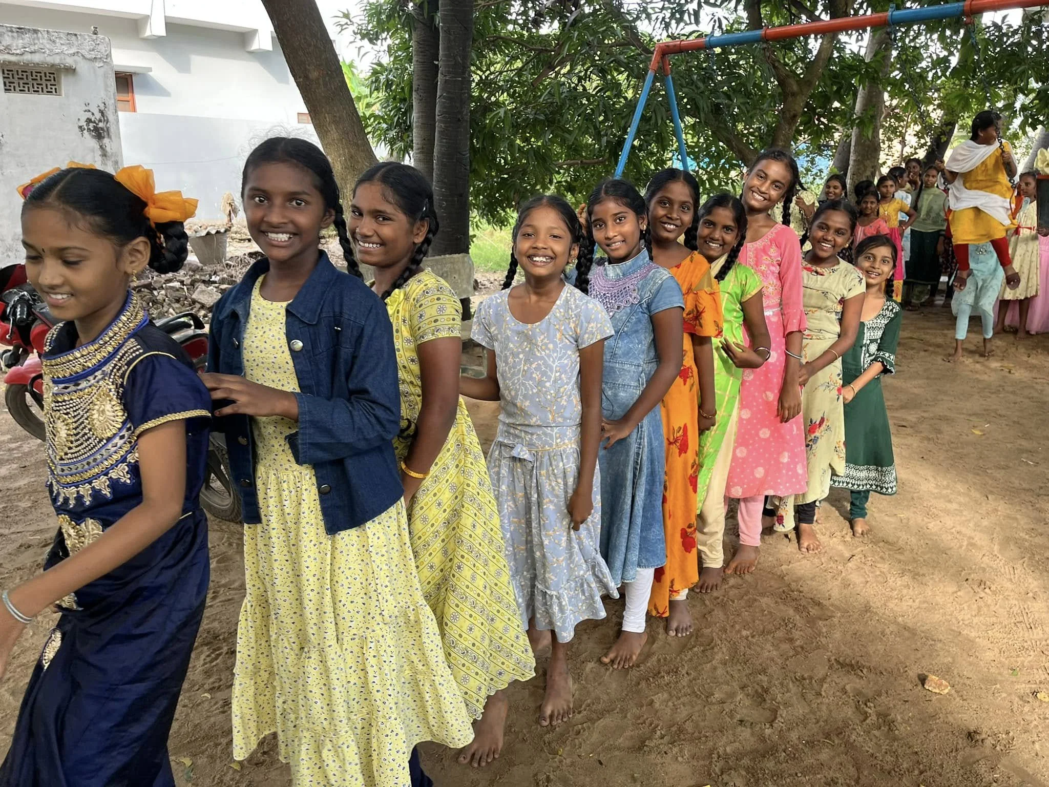 A group of smiling young girls standing in a line outdoors, dressed in colorful traditional clothing, with a playground and more children visible in the background.