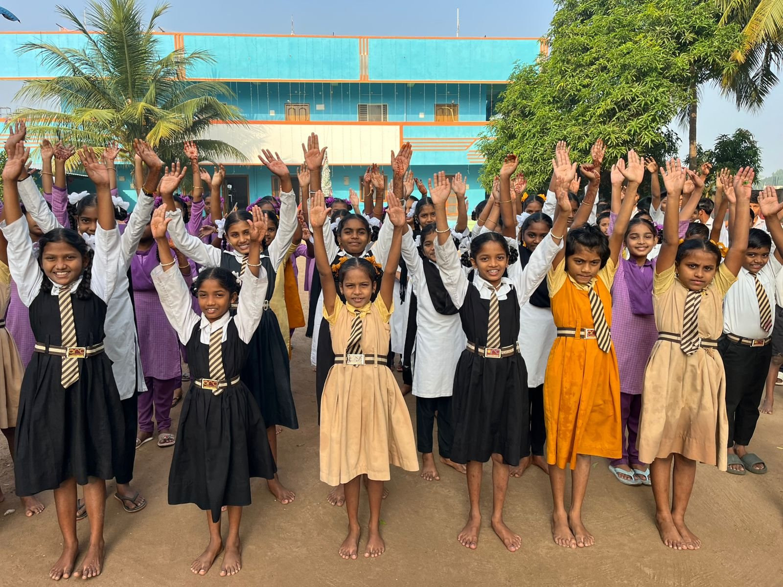 Group of school children standing outside, raising their hands, smiling, and wearing school uniforms in front of a school building and trees.