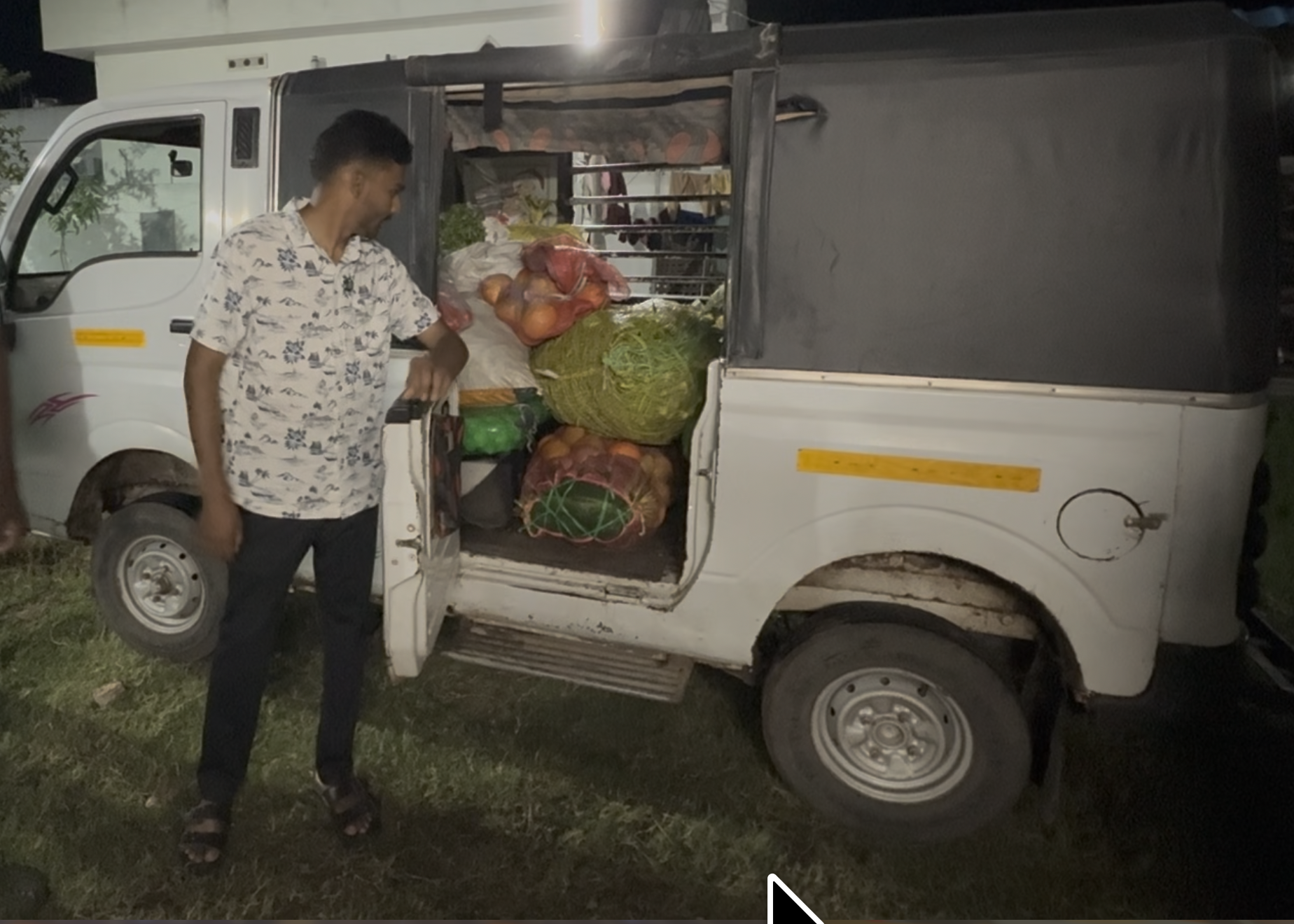 A man in a white floral shirt standing next to a small white truck with an open back, loaded with bags of vegetables including onions, cabbage, and other produce.