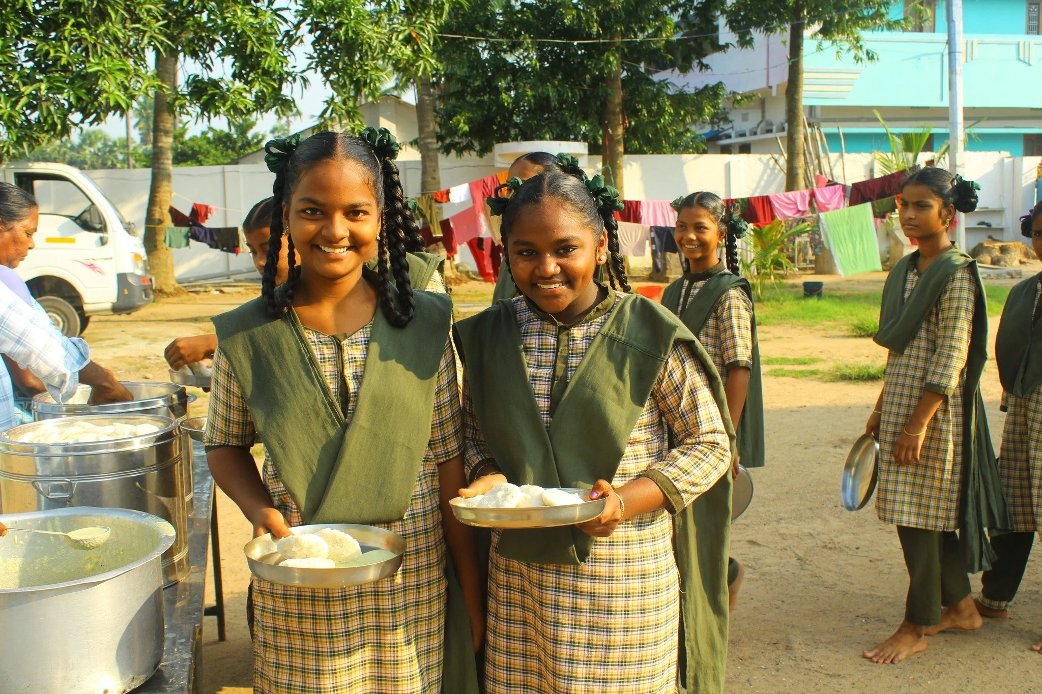 Girls in school uniforms smiling and holding plates of food outdoors with laundry hanging on a clothesline in the background.