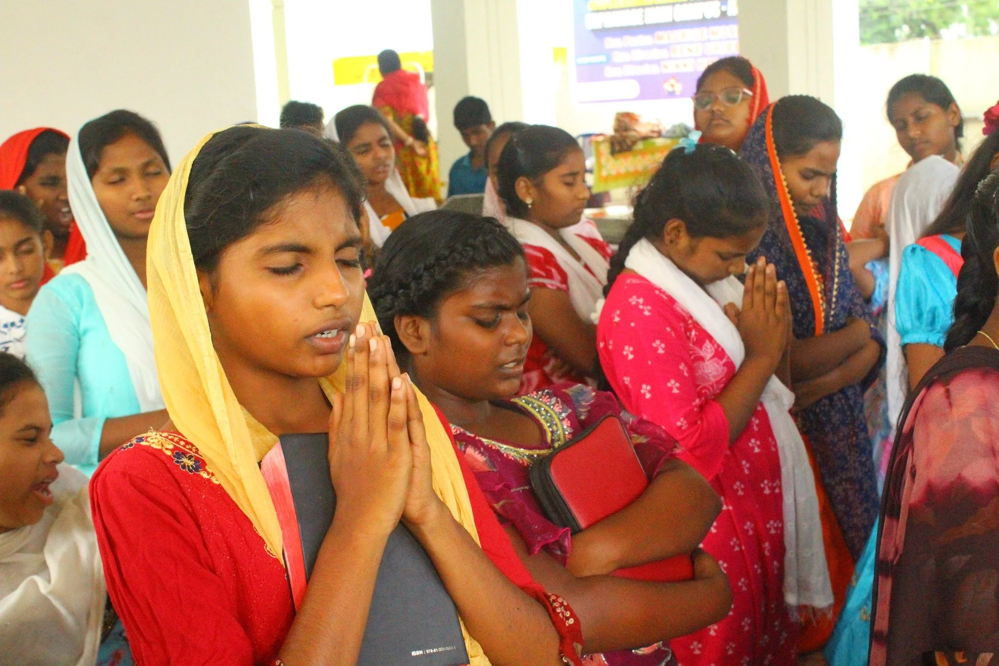 Group of young girls praying with hands clasped and eyes closed in a brightly lit prayer room.