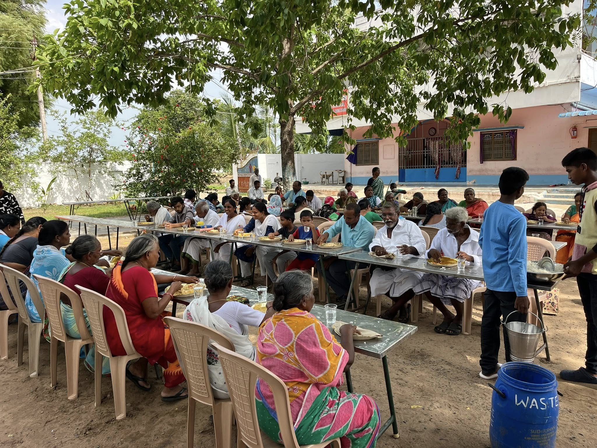 Outdoor gathering of people, mostly elderly, sitting at long tables eating food, under a large tree with a pink building in the background, in what appears to be a community event in India.