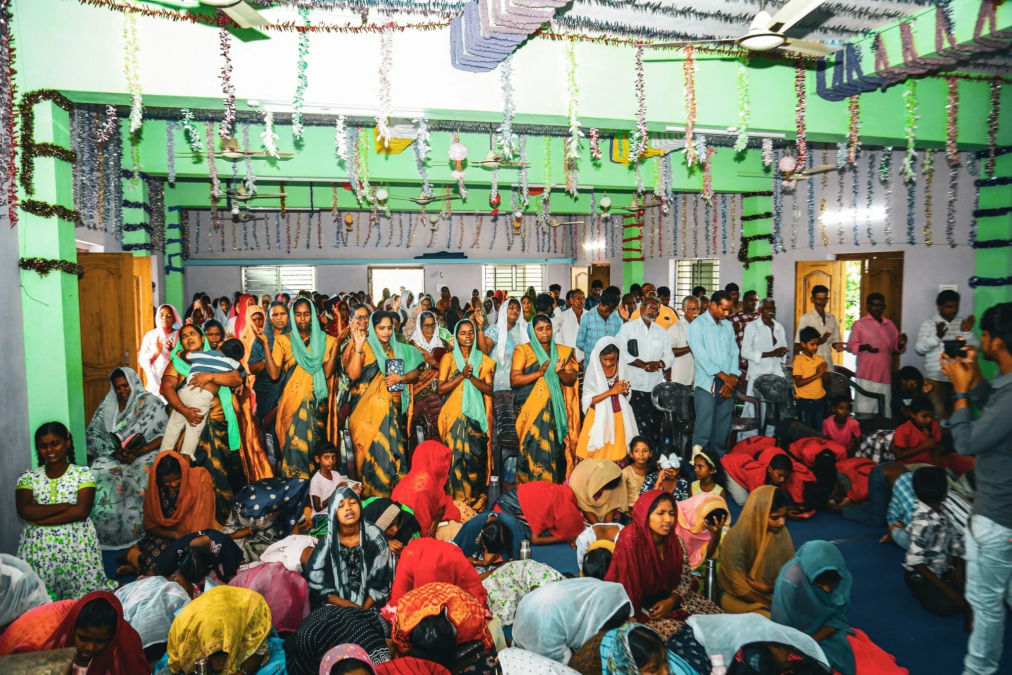 A large gathering of people, including men, women, and children, participating in a religious or spiritual ceremony inside a decorated hall. Many are kneeling or praying, with some standing, in colorful traditional attire. The hall is decorated with hanging streamers and garlands, and sunlight is coming through the open windows.