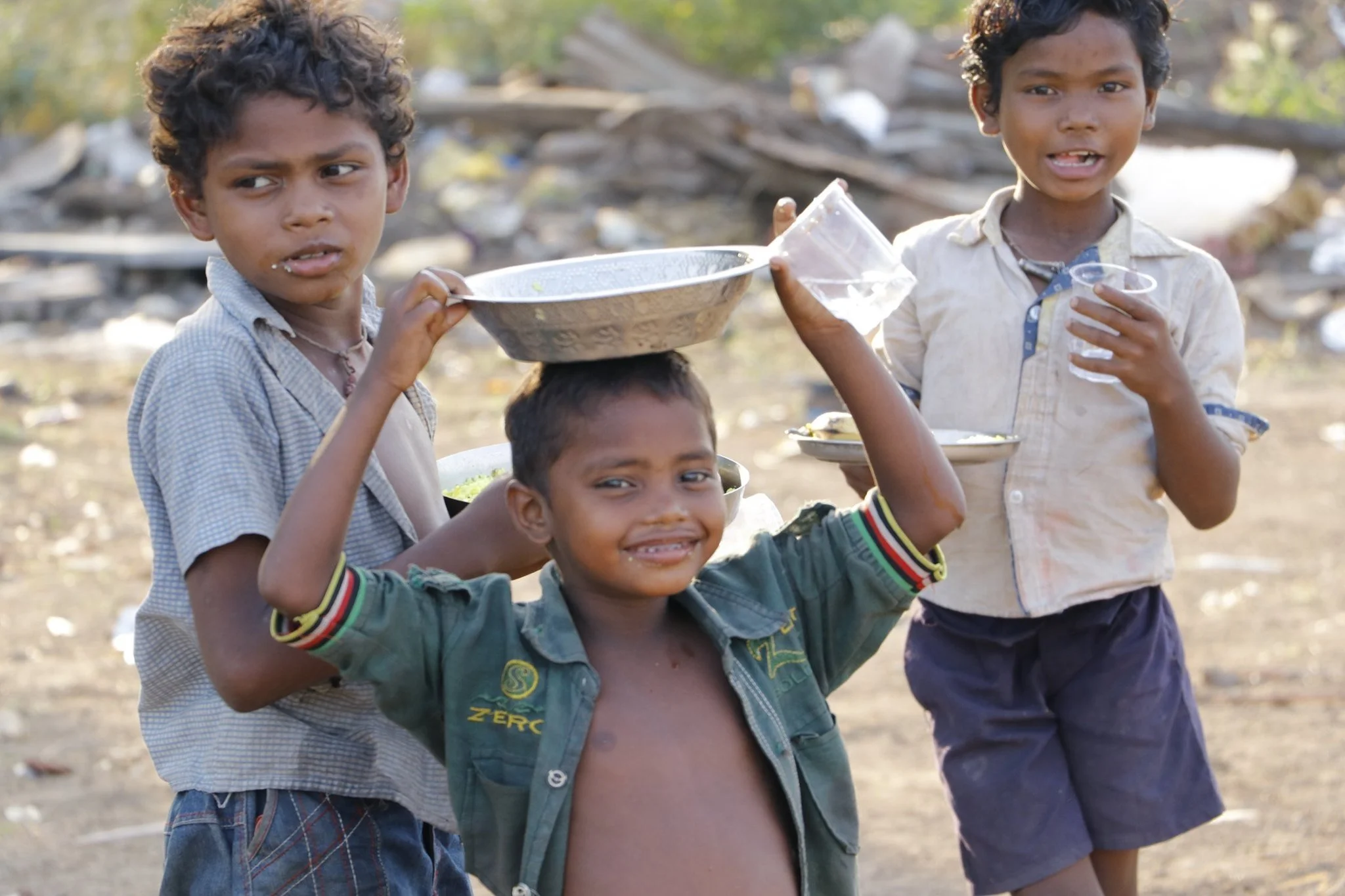 Four young children with dark curly hair and dark skin standing outdoors, holding food and drinks, on a dirt area with discarded trash and debris in the background.