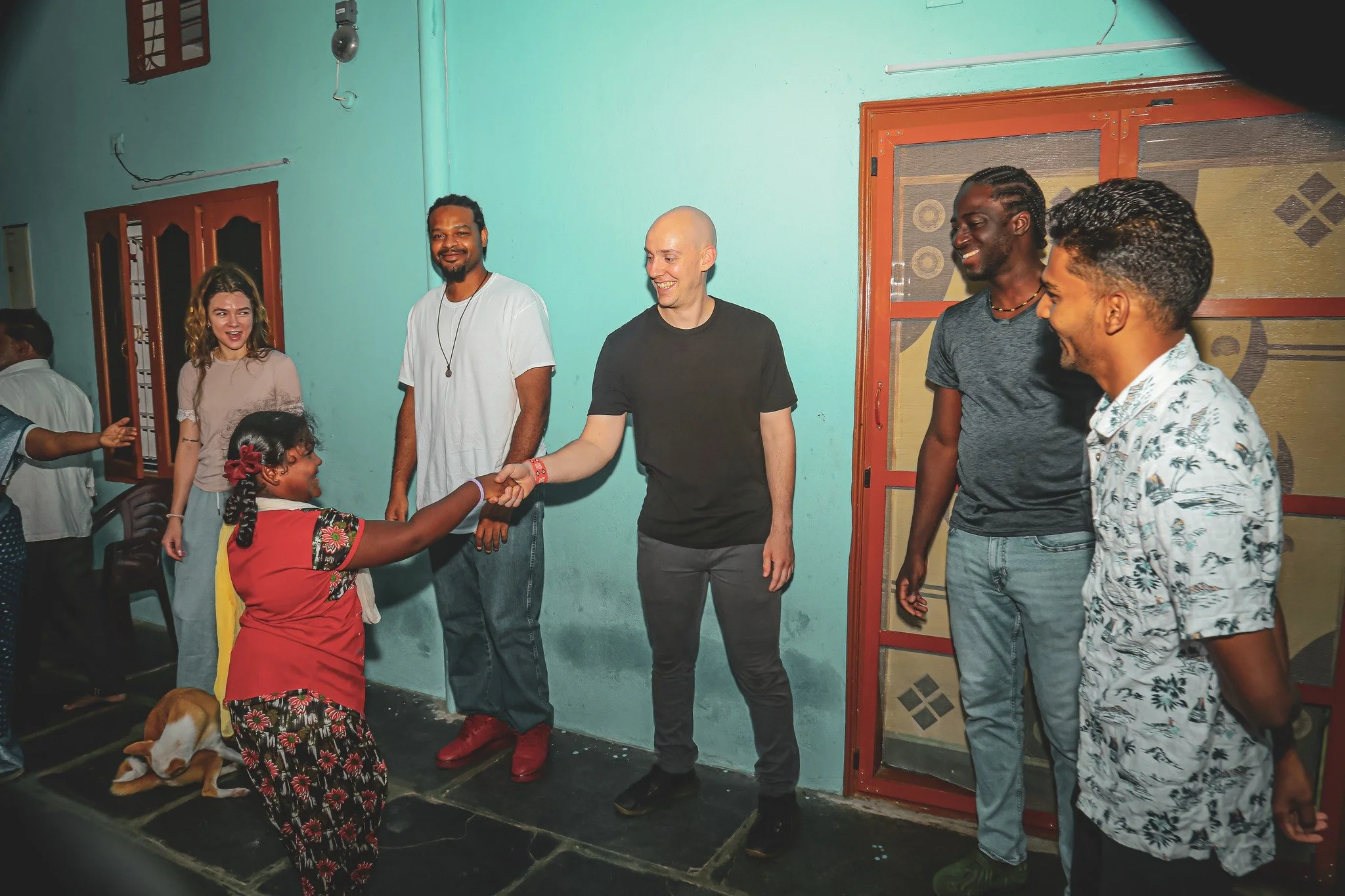 A group of people, including a young girl, are indoors. The girl, wearing a red dress with floral patterns, is shaking hands with a bald man in a black t-shirt. Three other men and a woman stand nearby, smiling and watching the interaction.