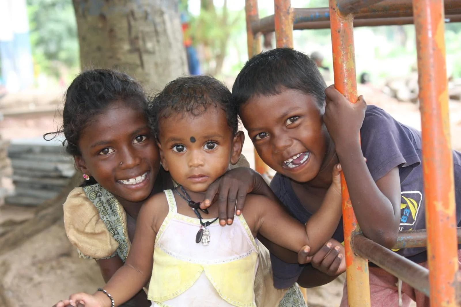 Three happy children, two boys and one girl, playing outdoors near a metal railing and a large tree. The children are smiling and looking at the camera.