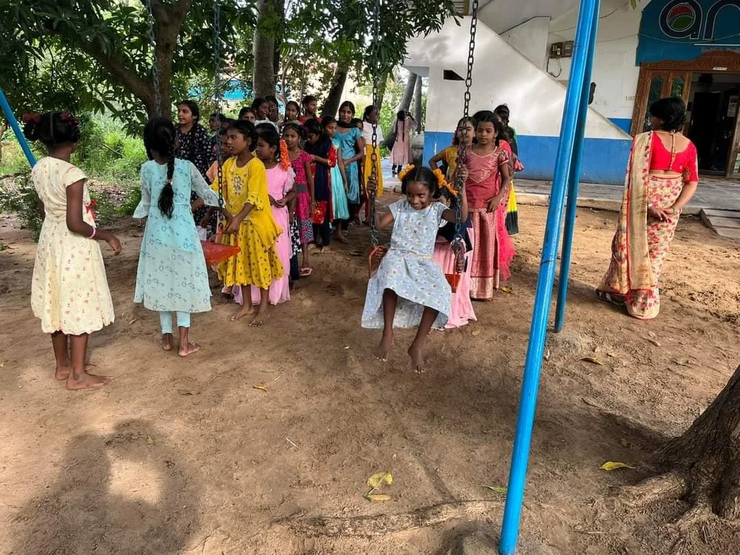 Girls in colorful dresses playing on a swing in a schoolyard with trees and a building in the background.
