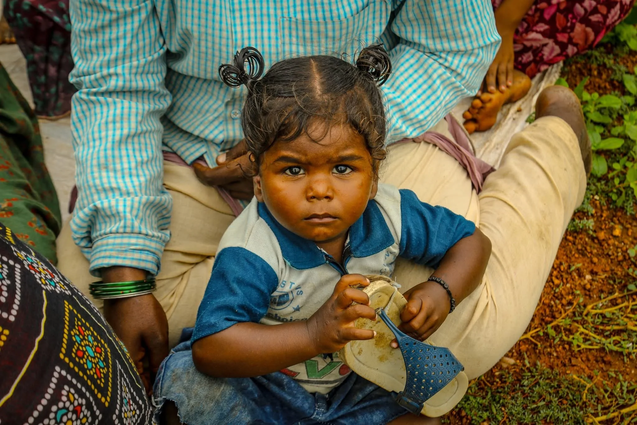A young girl with dark curly hair in pigtails, wearing a blue and gray shirt, sitting on the ground with a serious expression, holding a blue spatula. An adult is sitting behind her, with legs crossed and wearing beige pants and a blue striped shirt, on a grassy ground.