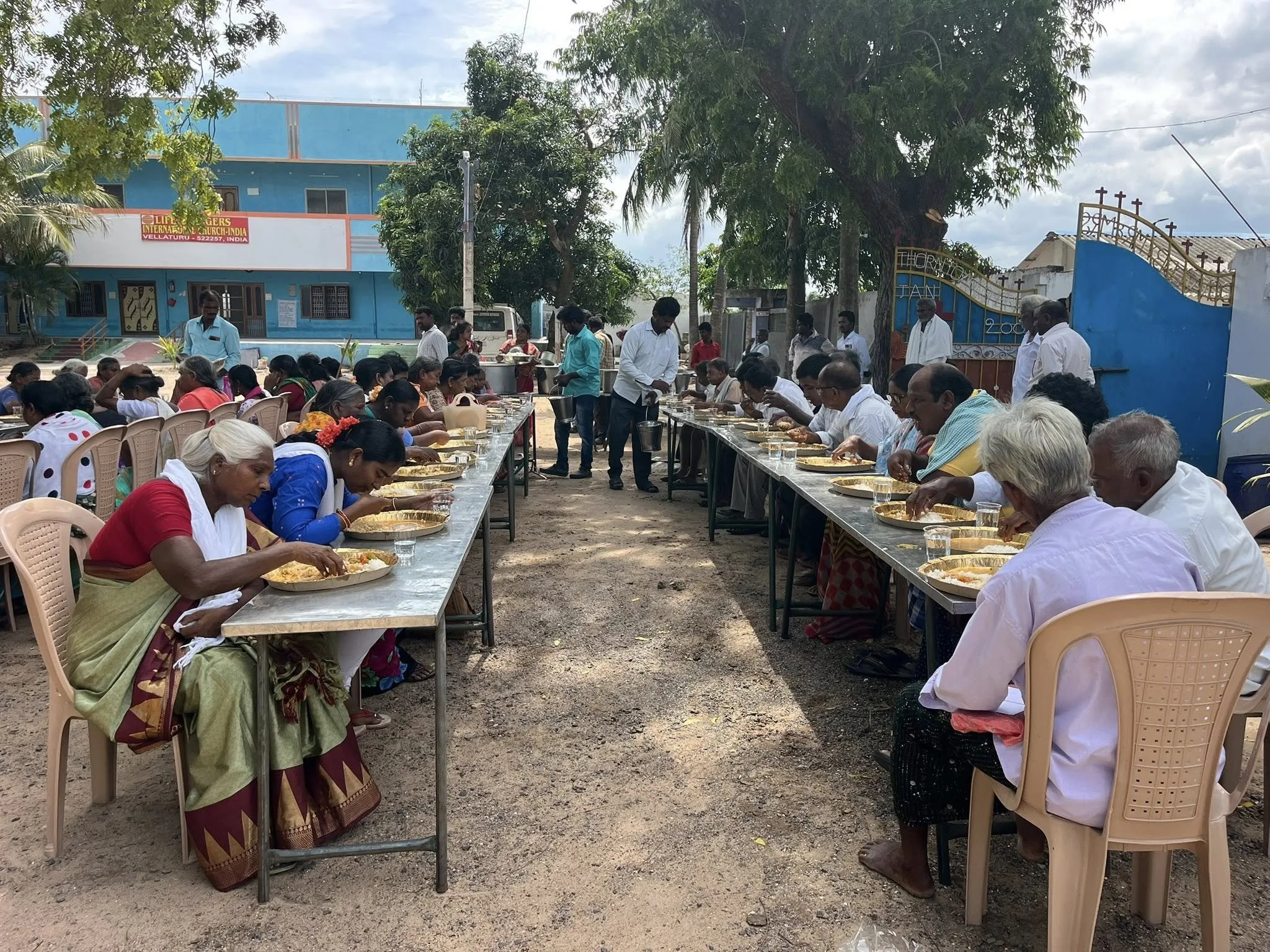People sitting at long tables outdoors, eating a meal, with a church building and trees in the background.