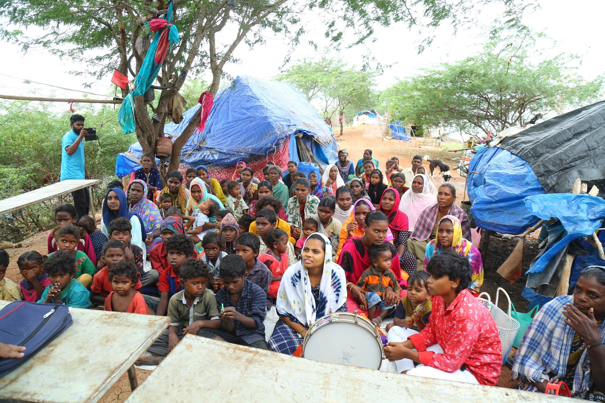 A large group of people, including women and children, gathered outdoors in front of makeshift tents covered with blue and black tarps. Some are sitting and some are standing, with a tree and sparse greenery in the background. A woman in red is playing a drum, and a man is taking a photo or video.