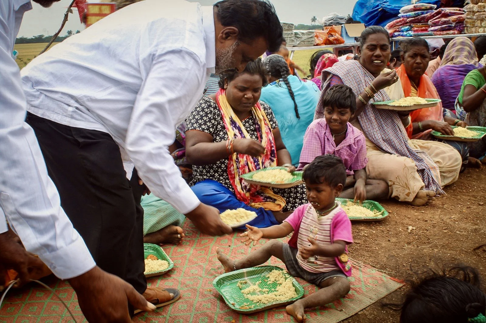 People sitting on the ground in a line, receiving food from a man. Several women and children hold plates of rice, and the children are barefoot and sitting on mats.