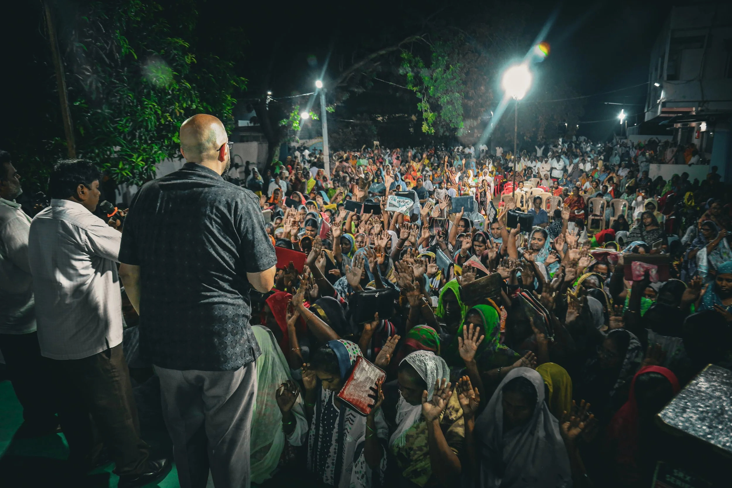 A crowd of people gathered outdoors at night, raising their hands, listening to a speaker on stage, with bright lights overhead illuminating the scene.